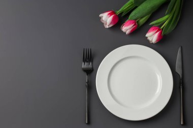 Yaratıcı masa, beyaz tabak, siyah çatal ve bıçak, koyu arkaplanda laleler. Sevgililer Günü, Düğün Günü, Doğum Günü, Kadınlar Günü ve Anneler Günü. Düz katman. Boşluğu kopyala.