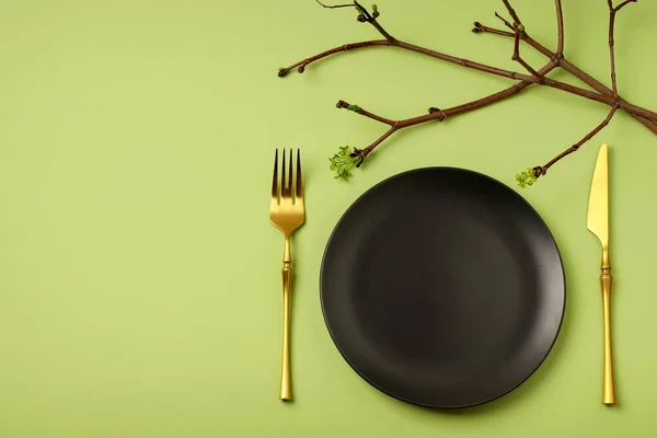 Black plate, gold table knife and fork on a green background. Spring setting of the festive table. Branches with leaves. An elegant dinner. Flat lay. Copy space