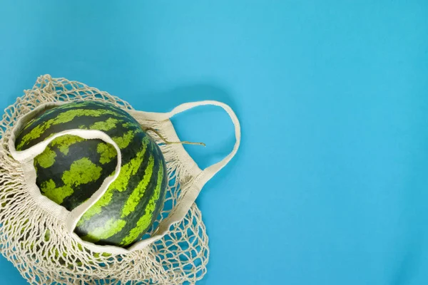 Healthy food delivery concept. Watermelon in a string bag on a blue background. Detox in a reusable eco mesh. Copy space