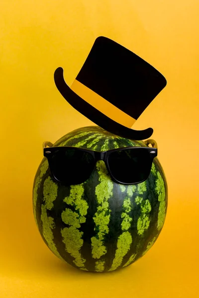 穿着黑色太阳镜 头戴黄色背景帽子的时髦西瓜 夏日水果的节日简约主义概念 纵向立场 图库图片