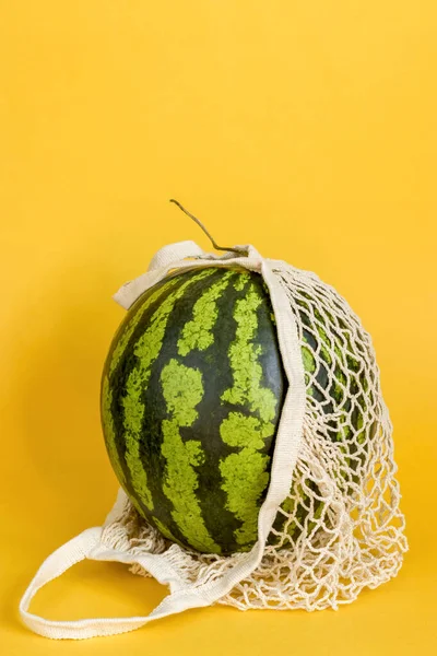 Watermelon in a string bag on a yellow background. Detox in a reusable eco mesh. Healthy food delivery concept. Copy space