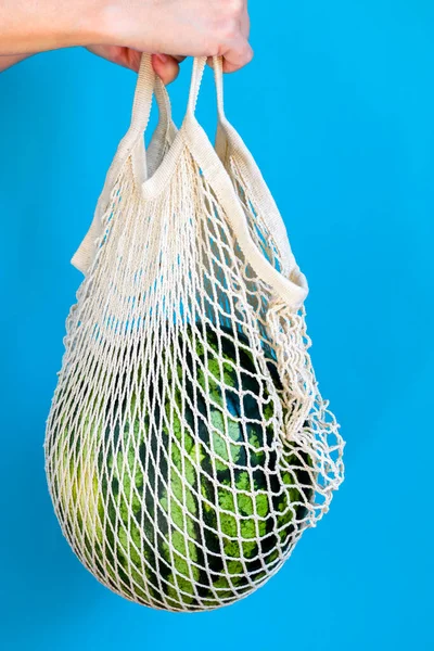 Hands hold a watermelon in a string bag on a blue background. Healthy food delivery concept. Detox in a reusable eco-mesh. Vertical position.