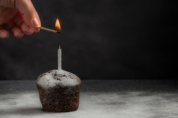 A female hand lights a candle on a delicious chocolate muffin with powdered sugar on a dark background. Birthday cake. Homemade baking. Festive concept. Low key. Copy space
