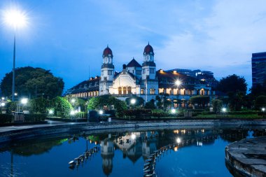 Semarang, Endonezya - 14 Aralık 2025: Lawang Sewu 'nun mavi saatte güzel bir uzun pozlama manzarası, tarihi bir dönüm noktası ve koloni binası aydınlatılmış fıskiye başlıkları ve park ışıkları ile ön plandaki bir gölete yansıdı.
