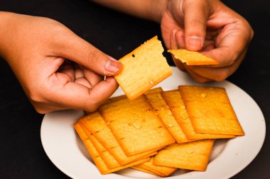 Beyaz Tabak Üzerine Çıtır Kraker Kıran Kişi, Fırın ve Mutfak Markası Tohumları ile Savory Golden Bisküvisi Kapalı, Siyah Masa Arkaplanı Kırıntılı Çıtır Atıştırmalık