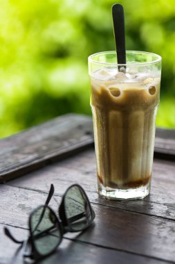 Tahta Masa üzerinde güneş gözlüklü buzlu kahve sütü, Doğal Yeşil Bokeh Arkaplanlı yaz içeceği konsepti, Gastronomi ve Açık Hava Kafe Yaşam Tarzı İçin Soğuk Latte