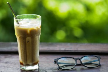 Buzlu sütlü buzlu kahve ve kırsal ahşap masa üzerinde güneş gözlüğü, Doğal Yeşil Bokeh Gastronomi ve Yaşam Tarzı Konsepti için arka planlı yaz içeceği