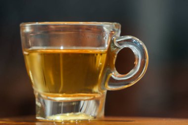Steaming Hot Black Tea in Small Glass Cup with Round Handle, Warm Golden Herbal Drink on Wooden Table, Professional Gastronomy Photography in Cozy Cafe Interior with Soft Bokeh Background