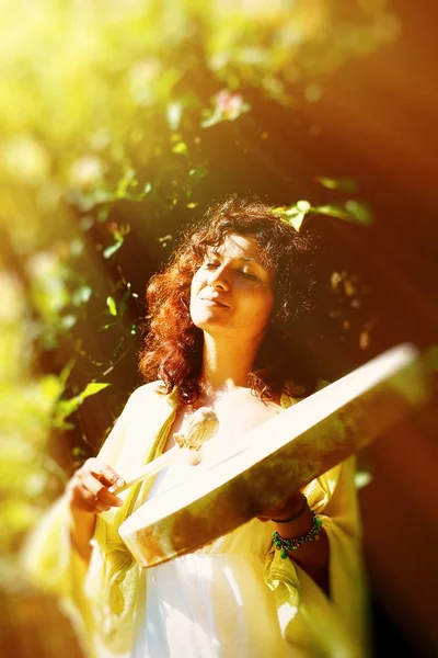 葉や花を背景にシャーマンのフレームドラムで遊ぶ美しいシャーマンの女の子。太陽光効果と夢のような効果. — ストック写真