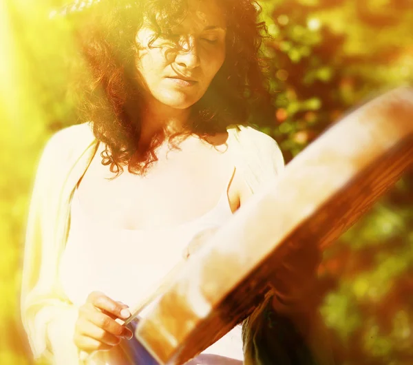 Bella ragazza sciamanica che gioca sul tamburo telaio sciamano su sfondo con foglie e fiori. effetto di luce solare e effetto di sfocatura sognante. — Foto Stock