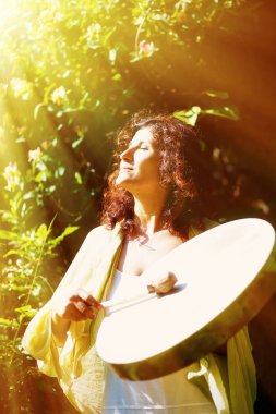 Şamanik güzel bir kız arka planda yapraklar ve çiçeklerle şaman çerçevesinde davul çalıyor. Güneş ışığı etkisi.