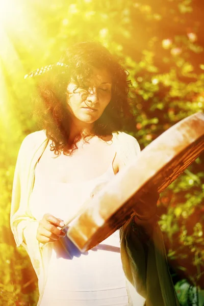 Hermosa chica chamánica jugando en tambor marco chamán en el fondo con hojas y flores. efecto de luz solar. — Foto de Stock
