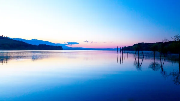 Gyönyörű táj, ahol egy tó és hegyek a háttérben, és a víz a fák. Kék és lila színű árnyalattól. — Stock Fotó