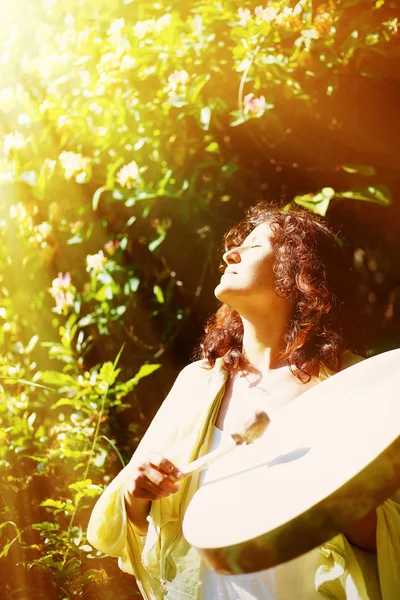 Hermosa chica chamánica jugando en tambor marco chamán en el fondo con hojas y flores. efecto de luz solar. — Foto de Stock