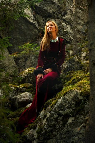 Una hermosa hada mujer con el pelo largo y rubio en un vestido histórico está sentada en medio de moos cubierto de rocas en el encantador paisaje forestal — Foto de Stock