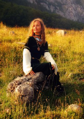 Mystical medieval maiden sitting on a wild meadow stone in a mountain meadow, looking up, with her face gently enlighted clipart