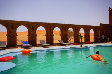 A beautiful moment of relaxation in a hotel swimming pool with ancient architecture amids moroccan desert dunes clipart