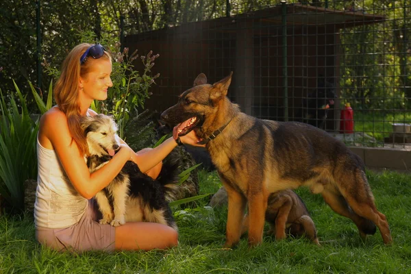 Joven hermosa mujer con el pelo rubio está sosteniendo amorosamente un perro callejero en sus brazos sentado en un jardín del patio trasero con hierba verde — Foto de Stock