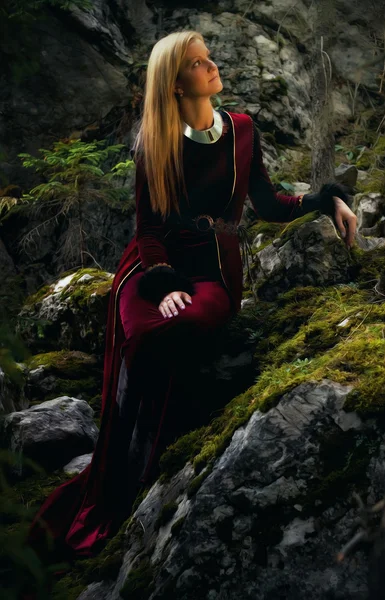 Hermosa hada mujer con el pelo largo y rubio en un vestido histórico está sentado en medio moos cubierto de rocas en el encantador paisaje forestal — Foto de Stock