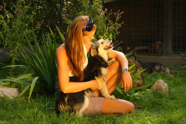 Joven hermosa mujer con el pelo rubio está sosteniendo amorosamente un perro callejero en sus brazos sentado en un jardín del patio trasero con hierba verde —  Fotos de Stock