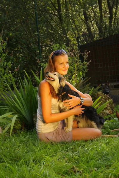 Joven hermosa mujer con el pelo rubio está sosteniendo amorosamente un perro callejero en sus brazos sentado en un jardín del patio trasero con hierba verde —  Fotos de Stock