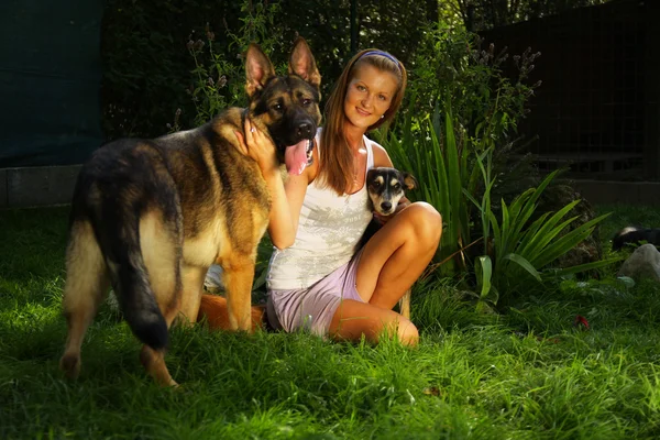 Joven hermosa mujer con el pelo rubio está sosteniendo amorosamente un perro callejero en sus brazos sentado en un jardín del patio trasero con hierba verde —  Fotos de Stock