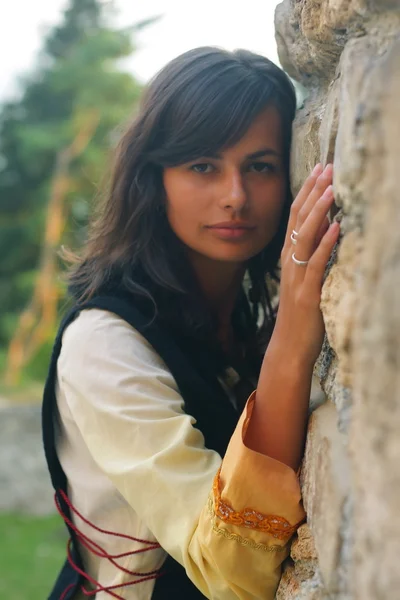 A beautiful young woman with long dark hair and a historical dress at a rough ancient stone wall — Stock Photo, Image