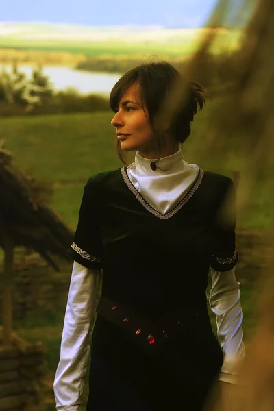 Uma bela jovem com cabelo escuro e um vestido histórico de veludo preto caminhando por uma paisagem aberta com um lago — Fotografia de Stock
