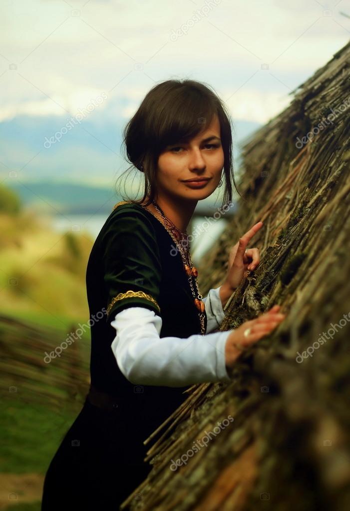 A beautiful young woman with dark hair, black velvet historical dress and a subtle smile is touching gently the rough surface of an ancient straw house roof