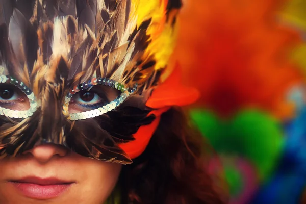 Mujer joven con una máscara de carnaval de plumas de colores sobre un fondo colorido brillante, contacto visual, maquillaje artista — Foto de Stock