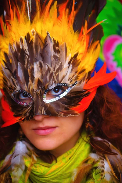 Mujer joven con una máscara de carnaval de plumas de colores sobre un fondo colorido brillante, contacto visual, maquillaje artista — Foto de Stock