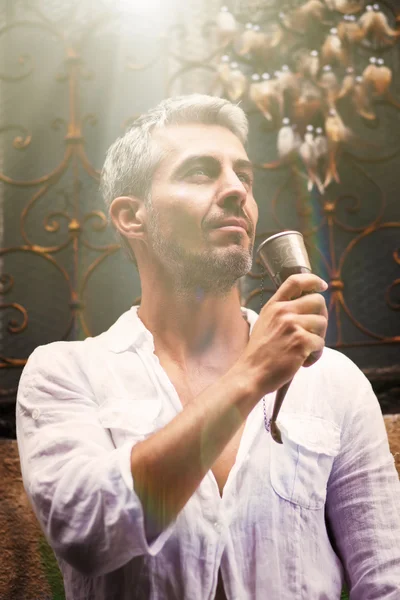 Sexi Man in a white shirt and medieval mead horn in hand. Toast concept. Ornamental window on background, and Dream Catcher. in sun light — Stock Photo, Image