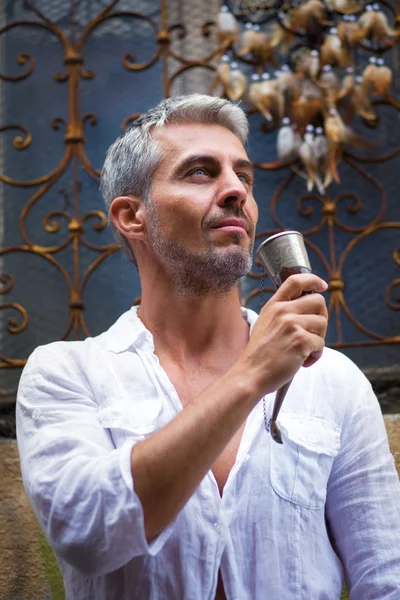 Sexi Man in a white shirt and medieval mead horn in hand. Toast concept. Ornamental window on background, and Dream Catcher. in sun light — Stock Photo, Image