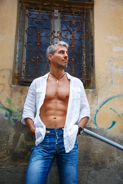 Sexy modelo masculino retrato de moda caliente en jeans elegantes y camisa con cuerpo muscular posando. Colgante de joyas de dientes de lobos . —  Fotos de Stock