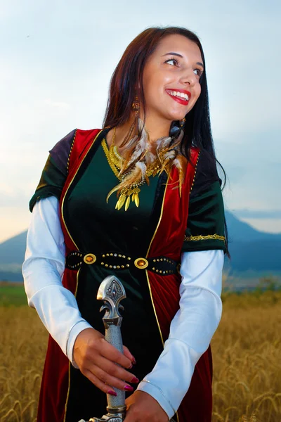 Smiling Young woman with ornamental dress and sword in hand  with sunset. Natural background.. — Stock Photo, Image