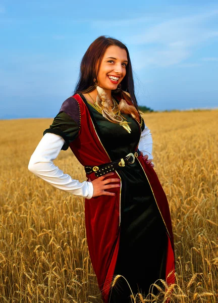 Smiling Young woman with medieval dress standing on a wheat field with sunset. Natural background. — Stock Photo, Image