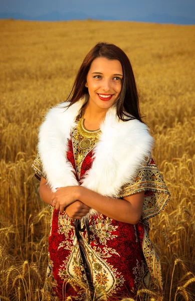 Young woman with ornamental dress and white fur standing on a wheat field with sunset. Natural background.. — Stock Photo, Image