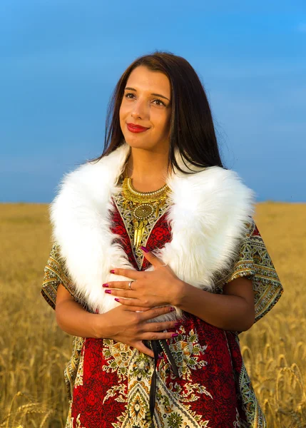 Young woman with ornamental dress and white fur standing on a wheat field with sunset. Natural background.. — Stock Photo, Image