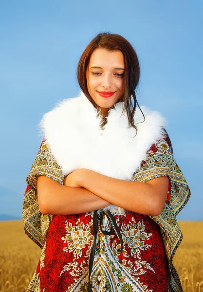 Young woman with ornamental dress and white fur standing on a wheat field with sunset. Natural background.. — Stockfoto