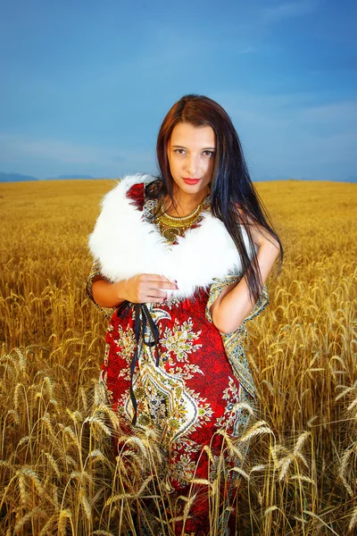 Smiling Young woman with ornamental dress and white fur standing on a wheat field with sunset. Natural background.. — Stock Photo, Image