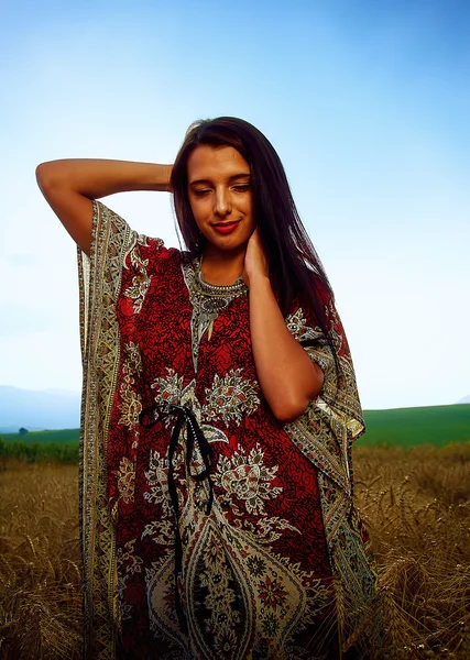 Jeune femme souriante avec robe ornementale debout sur un champ de blé avec coucher de soleil. Contexte naturel .. — Photo