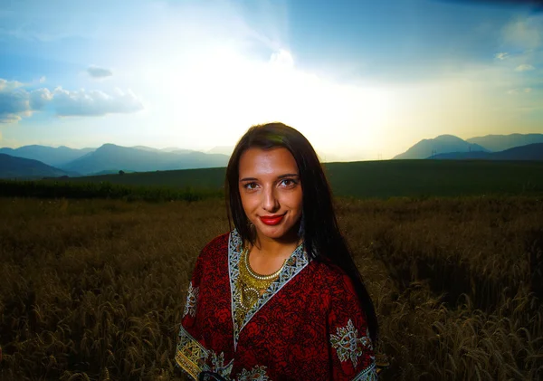 Jeune femme souriante avec robe ornementale debout sur un champ de blé avec coucher de soleil et ciel bleu. Contexte naturel .. — Photo