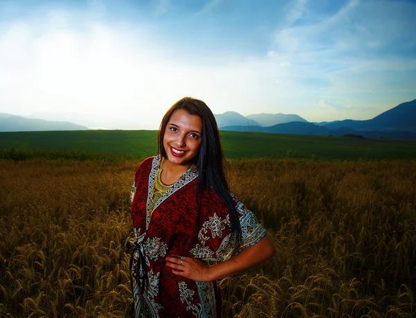 Jeune femme souriante avec robe ornementale debout sur un champ de blé avec coucher de soleil .. — Photo