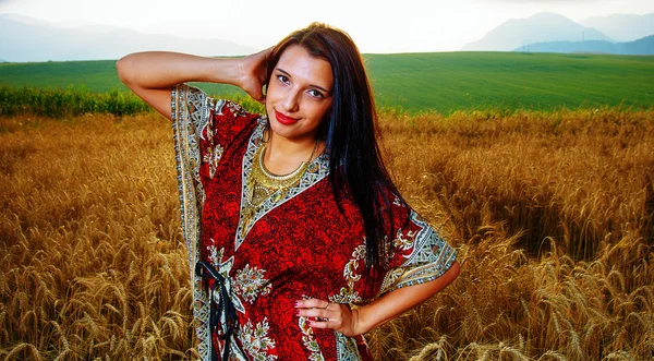 Jeune femme souriante avec robe ornementale debout sur un champ de blé avec coucher de soleil. Contexte naturel .. — Photo