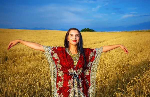 Leende ung kvinna med prydnads klänning stående på en veteåker med solnedgång. Naturlig bakgrund och blå himmel. — Stockfoto