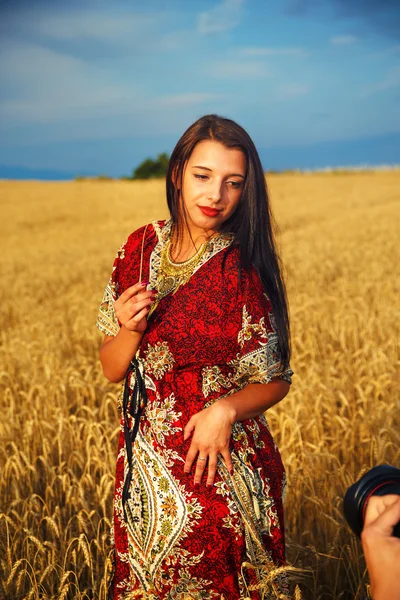 Leende ung kvinna med prydnads klänning stående på en veteåker med solnedgång. Och fotografen hand med kamera.. — Stockfoto