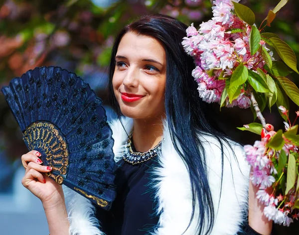 Hermosa mujer con flores, glamour de piel blanca y abanico negro en la mano, posando junto a flores mágicas de primavera rosa sakura. Fondo de flores — Foto de Stock