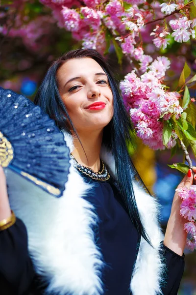 Beautiful Girl with flowers, glamour white fur and black fan in hand, posing next to blooming magical spring rosa sakura flowers. Flower background — Stock Photo, Image