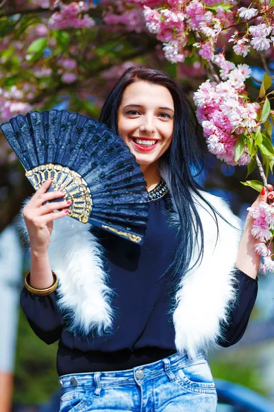 Beautiful Girl with flowers, glamour white fur and black fan in hand, posing next to blooming magical spring rosa sakura flowers. Flower background — Stock Photo, Image