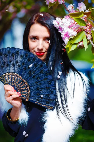 Beautiful Girl with flowers, glamour white fur and black fan in hand, posing next to blooming magical spring rosa sakura flowers. Flower background — Stock Photo, Image